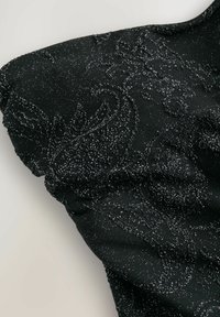 Černý top s texturovaným, lesklým vzorem a složitými designy. Materiál se zdá být měkký s mírně elastickým povrchem.