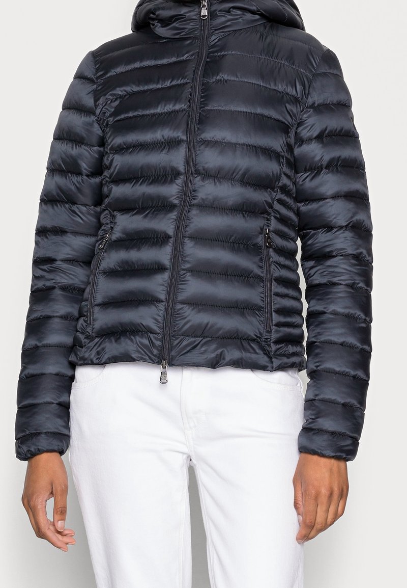 Personne portant une doudoune noire matelassée zippée avec capuche et un pantalon blanc, debout devant un fond clair uni.