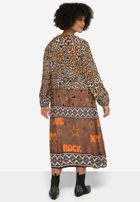 Robe longue à imprimé léopard en marron et orange, ornée de motifs étoilés, d'accentuations textuelles et d'une ceinture contrastante noire et blanche. Bottines noires à la cheville.