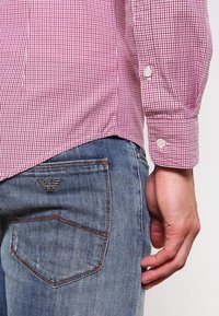 Muž v červené kostkované košili s dlouhým rukávem, zasunuté do modrých džínů, s viditelným logem Emporio Armani na zadní kapse.