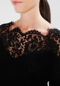 Schwarzes Oberteil mit einem Spitzenkragen mit gewelltem Rand und strukturierter Stoffoberfläche. Die Spitze zeigt filigrane Blumenmuster, die im Kontrast zum weichen Hauptstoff stehen.