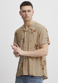 Camisa de manga corta beige con un patrón floral marrón, fabricada en tela ligera. Cuenta con un cuello muescado y cierre de botones.