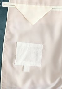 Producto textil con un acabado satinado beige, que presenta una solapa triangular y un bolsillo de fieltro blanco. Se aprecian una textura suave y bordes limpios.