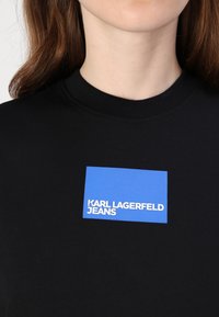 Černá mikina se skládá z obdélníkové modré nášivky s bílým textem "KARL LAGERFELD JEANS." Hladký povrch a podrobný žebrovaný límec.