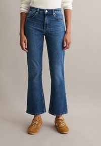 Person wearing mid-rise, slightly flared blue jeans with rolled cuffs and brown suede sneakers, standing against a plain neutral background.