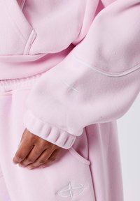 Haut à capuche rose avec un tissu texturé, des poignets côtelés et des accents brodés sur la manche et la poche pour un détail supplémentaire.