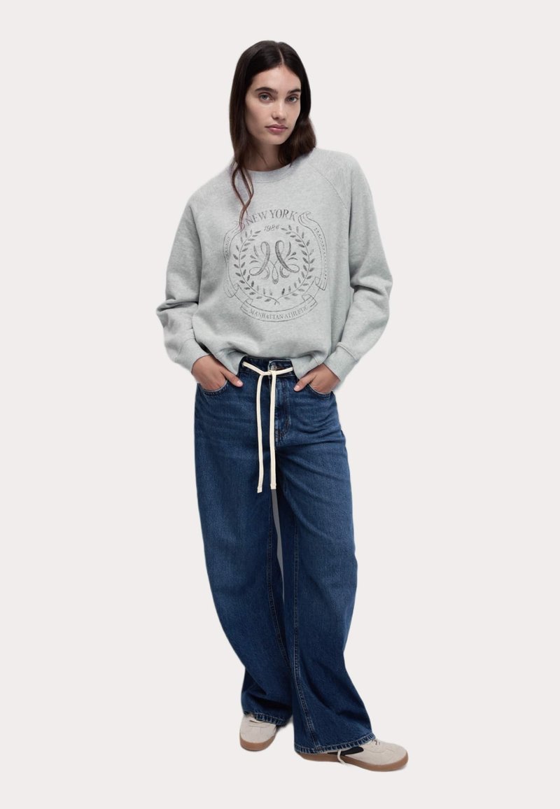 Felpa grigia con stampa a logo circolare, abbinata a jeans in denim blu larghi e sneakers grigie con suola bianca. Vestibilità rilassata, stile casual.