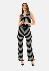 Combinaison à motifs noir et blanc avec un haut ajusté sans manches et un pantalon évasé, dotée d'une fermeture à nouer et d'une texture lisse. Talons noirs.