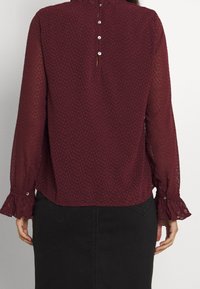 Bordeauxrote Bluse mit strukturiertem Stoff, gerafftem Ausschnitt und langen, transparenten Ärmeln. Mit Knopfverschluss und gerüschten Manschetten.