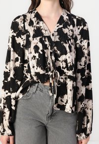 Schwarz-weiße gemusterte Bluse mit langen Ärmeln, elastischem Bund und einem Bindeband vorne, kombiniert mit grauen, hoch taillierten Hosen.