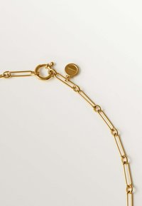 Collar de cadena de metal dorado que presenta eslabones alargados, un cierre circular y una pequeña etiqueta redonda con detalles en relieve.