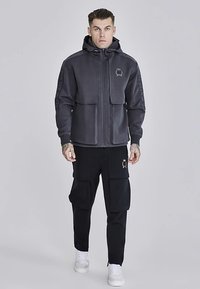 Sivi pulover z zadrgo in visokim ovratnikom ter dvema sprednjima žepoma, kombiniran s črnimi cargo hlačami z več žepi in belimi supergami.