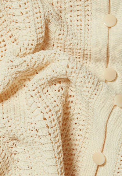 Pull en crochet crème clair avec un motif de tricot texturé et de grands boutons beiges sur le devant. Matière douce et confortable.