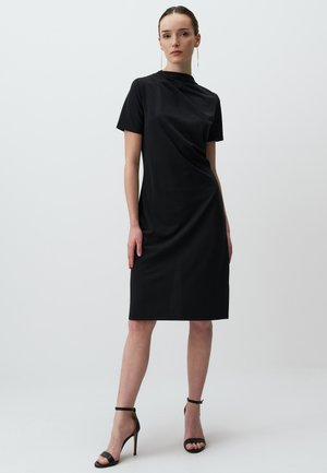 Abito nero a maniche corte con collo alto e design drappeggiato. Indossato con tacchi neri con cinturini, presenta una siluetta aderente e un taglio fino al ginocchio.