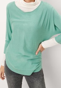 Pull vert d'eau clair avec un large décolleté, manches longues et coupe ample, superposé à un col roulé blanc. Texture lisse et ourlet arrondi.