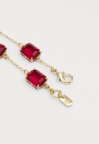 Cadena en tono dorado con dos grandes joyas rectangulares rojas con bordes biselados, acabada con un broche de langosta y una pequeña etiqueta.
