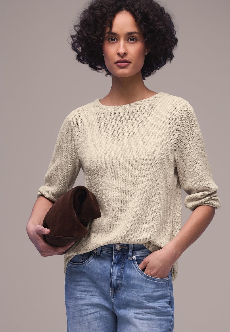 Kvinde med krøllet hår iført beige strikket sweater og blå jeans, der holder en foldet brun ruskindsjakke i den ene hånd og har den anden hånd i lommen.