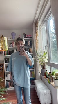 Světle modré oversized tričko s černými akcenty, s krátkými rukávy a zaobleným lemem, s textem na přední straně. Nosené se světlými džíny.