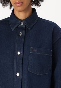 Tumed sinine denimsärk klassikalise kraega, suurte nööpidega ja rinnataskuga, millel on väike punane silt. Tekstureeritud kangas.