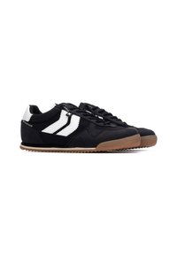 Zapatilla negra con rayas blancas y parte superior sintética. Presenta una suela de goma y cordones negros, con una textura suave y punta redondeada.