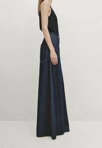 Femme portant un haut sans manches noir et un jean en denim foncé à taille haute et jambes larges, se tenant de profil contre un fond uni.