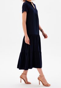 Kvinna i en marinblå, lager-på-lager midi-klänning med korta ärmar och nude högklackade ankelsandaler, stående mot en vit bakgrund.
