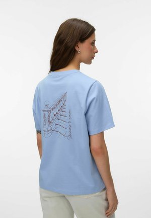 Kobieta o długich brązowych włosach nosi jasnoniebieską koszulkę z rysunkiem schodów i napisem "Schody do nieba" z tyłu, w połączeniu z jasnobeżowymi spodniami.