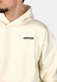 Felpa color crema realizzata in morbido tessuto, dotata di tasca a marsupio frontale e logo "CRYSTAL" in bold nero.