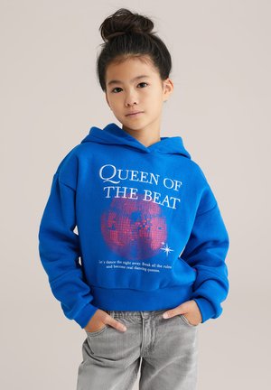 Blå hoodie med texten "QUEEN OF THE BEAT", en discokula grafik och ribbade muddkanter. I kombination med ljusgrå jeans.