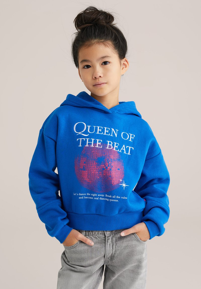 Modra kapuca z napisom "KRALJICA BEATA", grafiko disco krogle in z ribastimi manšetami. V kombinaciji z svetlo sivo denim hlačami.