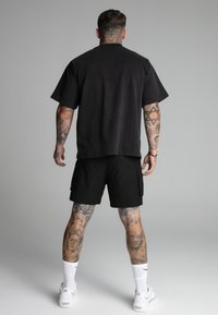 Crna oversized majica, pamučni materijal, kratki rukavi, u kombinaciji s crnim cargo šortsevima, bijelim tenisicama, vidljivim tetovažama na nogama.