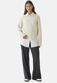 Lys beige chunky strikket sweater lagd over en hvid stribet skjorte med knapper, parret med mørkegrå brede bukser og casual sneakers.