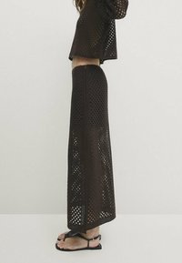 Dunkelbraunes, zweiteiliges Strickoutfit mit offenem Strickmuster. Das Oberteil ist leicht bauchfrei, der lange Rock hat seitliche Schlitze. Schwarze Sandalen.