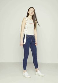 Top branco de alças com texto vermelho, jeans azuis, ténis brancos e uma mochila branca com uma alça studs. O modelo está em um fundo neutro.