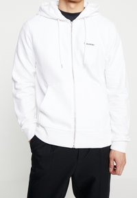 Man som bär en vit zip-up huvtröja med fickor fram och svarta byxor, stående med händerna i fickorna mot en vit bakgrund.