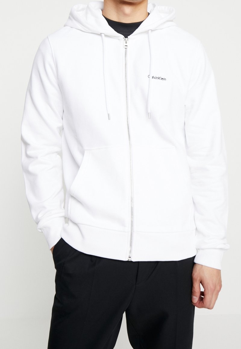 Man som bär en vit zip-up huvtröja med fickor fram och svarta byxor, stående med händerna i fickorna mot en vit bakgrund.