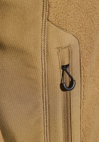 Beige fleecejacka med ribbat tyg, utrustad med en dragkedjeficka med en svart dragkedja, samt synliga sömdetaljer.