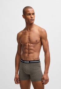 Boxer briefs in black and cream pattern, featuring a black waistband with "BOSS" logo. Fabric appears soft and stretchy. Model has a muscular build.