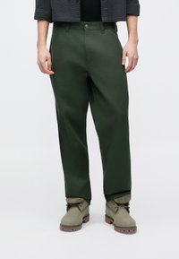 Σκούρο πράσινο παντελόνι με ψηλή μέση και άνετη γραμμή, με κούμπωμα με κουμπιά στο μπροστινό μέρος και πλαϊνές τσέπες, φορεμένο με γκρι μπότες.