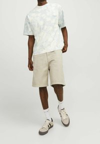 Muže v světlých béžových šortkách, košili s krátkým rukávem ve světlém vzoru, bílých ponožkách do poloviny lýtek a béžových teniskách s černými pruhy.