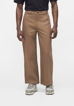 Mann trägt braune Hose mit weitem Bein, schwarzes T-Shirt und graue Wanderschuhe, steht vor weißem Hintergrund.