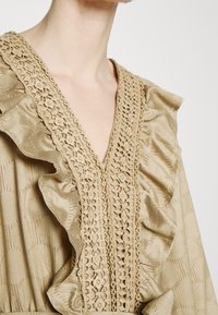 Robe beige avec un décolleté en V ornée de dentelle intricate et d'accents froncés, fabriquée à partir d'un tissu lisse et texturé.
