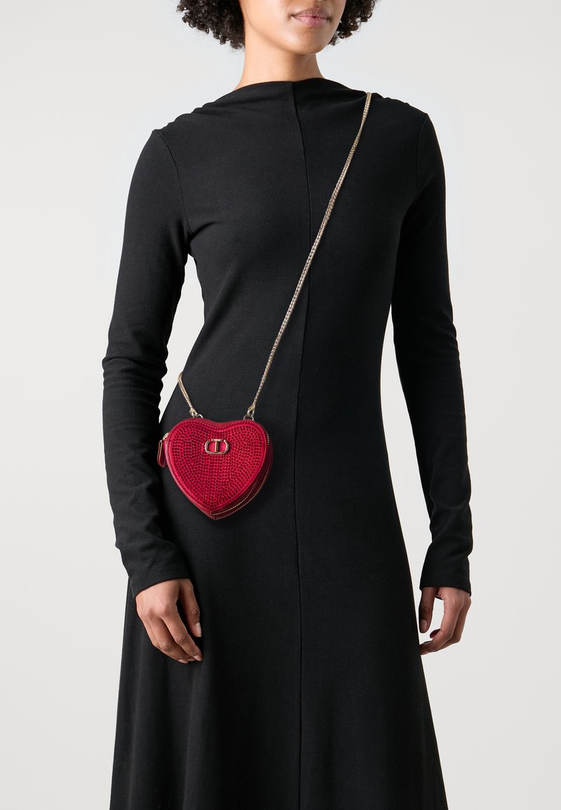 Donna che indossa un lungo abito nero con una piccola borsa a tracolla a forma di cuore rosso con una tracolla in oro.