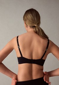 Naine näht tagant, kandes musta rinnahoidjat reguleeritavate rihmadega ning tema juuksed on madalasse hobusesaba seotud, taust on ühevärviline.