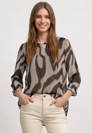 Jeune femme souriante, portant un chemisier à manches longues taupe et noir à motifs avec un pantalon beige, mains dans les poches, sur un fond uni.