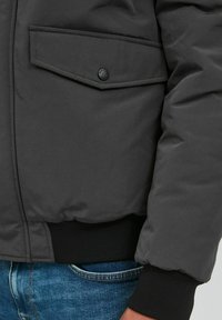 Gray insulated jacket with a textured finish, featuring a flap pocket, ribbed black cuffs, and a sleek, rounded silhouette.