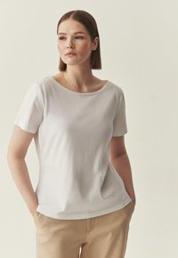 T-shirt blanc à manches courtes avec un col arrondi et une texture douce, présentant une coupe décontractée. Associé à un pantalon beige.