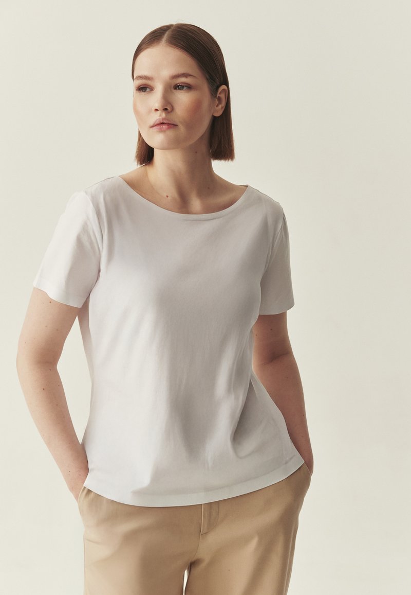 T-shirt blanc à manches courtes avec un col arrondi et une texture douce, présentant une coupe décontractée. Associé à un pantalon beige.