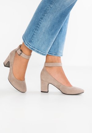 Pieds de femme portant des escarpins à talons blocs beige en suède avec brides à la cheville, associés à un jean bleu clair raccourci sur fond blanc.