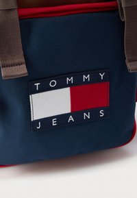 Geantă din pânză albastru marin cu bretele maro pentru umăr. Are o etichetă dreptunghiulară cu sigla „TOMMY JEANS”, cu accente roșii și albe.
